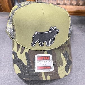May include: A green and camouflage trucker hat with a black pig silhouette patch. The hat features a foam front and a durable snap closure. The tag reads "World Famous Foam Front" and "Trucker".