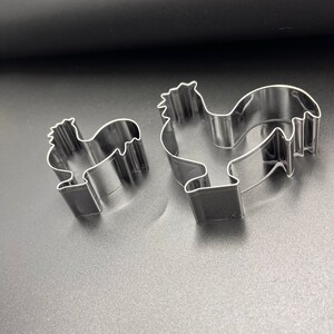 May include: Two stainless steel rooster-shaped cookie cutters. The cutters are in different sizes, with a detailed outline of a rooster's body, including a comb, beak, and tail feathers. The cookie cutters are on a dark gray surface.