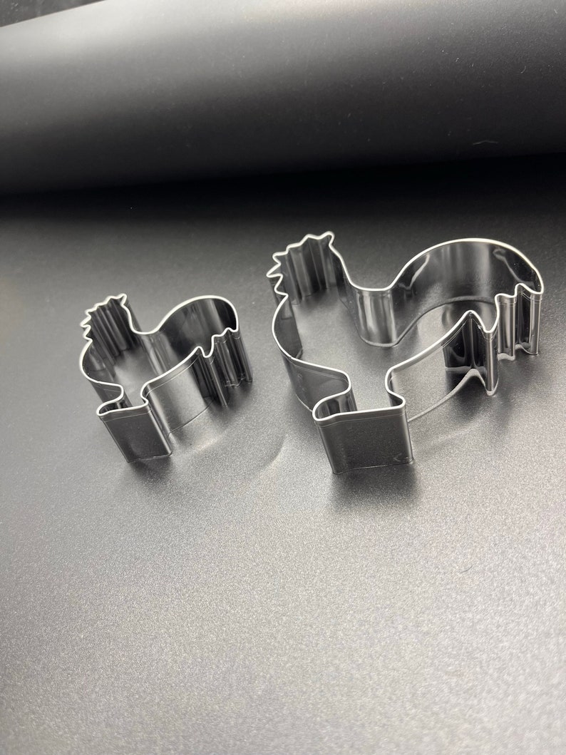 May include: Two stainless steel rooster-shaped cookie cutters. The cutters are in different sizes, with a detailed outline of a rooster's body, including a comb, beak, and tail feathers. The cookie cutters are on a dark gray surface.