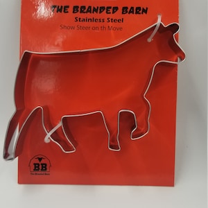 May include: A stainless steel cow-shaped cookie cutter on a red card. The card reads "THE BRANDED BARN" and "Show Steer on th Move". The cookie cutter is made of shiny, silver metal.