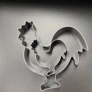 May include: Three stainless steel rooster-shaped cookie cutters. The cookie cutters are nested, with one slightly smaller than the other. The rooster's comb, wattle, and tail feathers are clearly defined. The cookie cutters are on a dark gray surface.