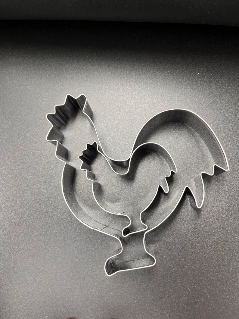 May include: Three stainless steel rooster-shaped cookie cutters. The cookie cutters are nested, with one slightly smaller than the other. The rooster's comb, wattle, and tail feathers are clearly defined. The cookie cutters are on a dark gray surface.