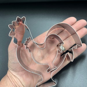 May include: A set of three stainless steel rooster-shaped cookie cutters. The cutters are nested inside each other, with the largest measuring approximately 6 inches in length. The design features a detailed comb, wattle, and tail feathers.