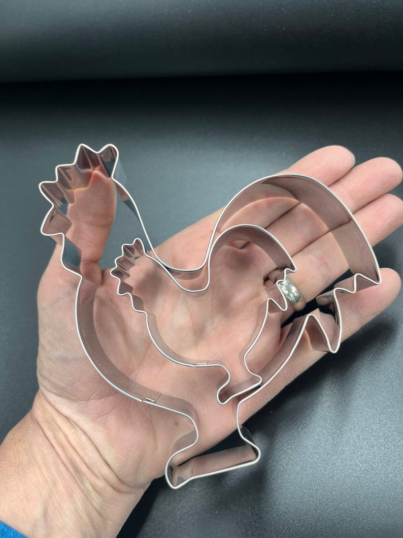 May include: A set of three stainless steel rooster-shaped cookie cutters. The cutters are nested inside each other, with the largest measuring approximately 6 inches in length. The design features a detailed comb, wattle, and tail feathers.