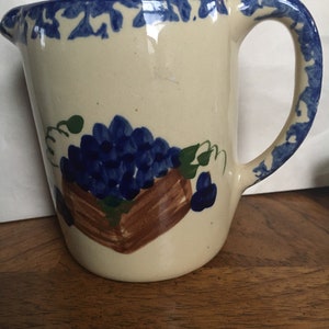 May include: A white ceramic pitcher with a blue and white floral design. The pitcher has a handle and a spout. The floral design features a bunch of blue grapes in a brown basket.