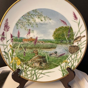 May include: A white porcelain plate with a gold rim features a painted scene of a duck family with ducklings in a grassy marsh. The scene includes a small bridge, a house, and a bird's nest with a bird and chicks.