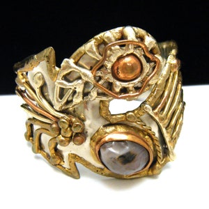 Puede incluir: Un brazalete de plata, oro y cobre con una piedra gris engastada en el centro. El brazalete está intrincadamente diseñado con un motivo floral y un patrón geométrico.