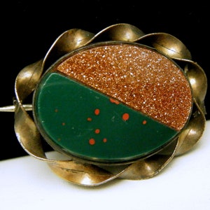 Puede incluir: Un broche vintage con una piedra verde y marr&oacute;n engastada en un marco de metal retorcido plateado.