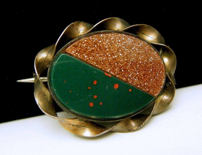 Puede incluir: Un broche vintage con una piedra verde y marr&oacute;n engastada en un marco de metal retorcido plateado.