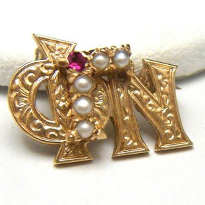 May include: Gold-toned sorority pin with the letters 'ΦΝ' and a red stone surrounded by pearls.