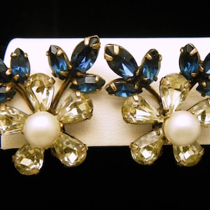 Op de afbeelding: Een paar gouden oorbellen met blauwe en heldere strass stenen in een bloemvormig ontwerp. Elke oorbel heeft een parel in het midden van de bloem.