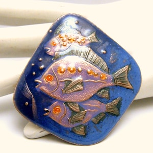 Puede incluir: Un pin de esmalte cuadrado con fondo azul y tres peces en tonos de morado, naranja y verde. Los peces tienen detalles dorados y están dispuestos en capas.