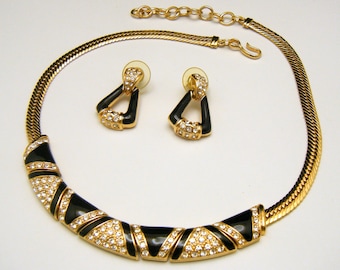 May include: A gold-toned necklace and earring set. The necklace features a black enamel design with clear crystal accents. The earrings are triangular with black enamel and crystal details. The set is displayed on a white background.