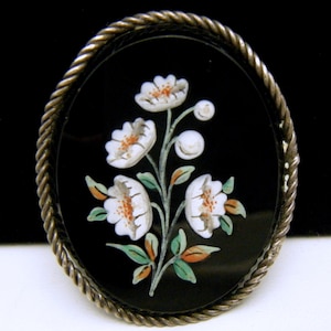 May include: An oval-shaped brooch with a black background and a white floral design. The brooch is surrounded by a silver rope-like border.