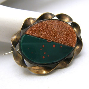 Puede incluir: Un broche vintage con un engaste de metal dorado y una piedra verde y marr&oacute;n. La piedra est&aacute; dividida en dos, una mitad es un marr&oacute;n brillante y la otra mitad es un verde con motas rojas.