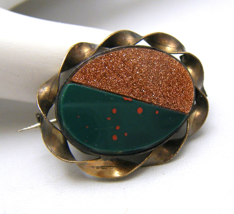 Puede incluir: Un broche vintage con un engaste de metal dorado y una piedra verde y marr&oacute;n. La piedra est&aacute; dividida en dos, una mitad es un marr&oacute;n brillante y la otra mitad es un verde con motas rojas.
