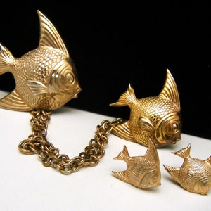 Puede incluir: Un broche de pescado dorado con una cadena a juego y un par de pendientes. Los peces están detallados con escamas texturizadas y tienen un diseño realista.