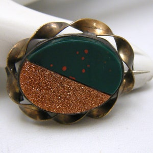 Puede incluir: Un broche vintage con una piedra de color verde y cobre engastada en un marco de lat&oacute;n. La piedra est&aacute; dividida en dos, un lado verde oscuro con motas naranjas y el otro lado un brillo de color cobre.
