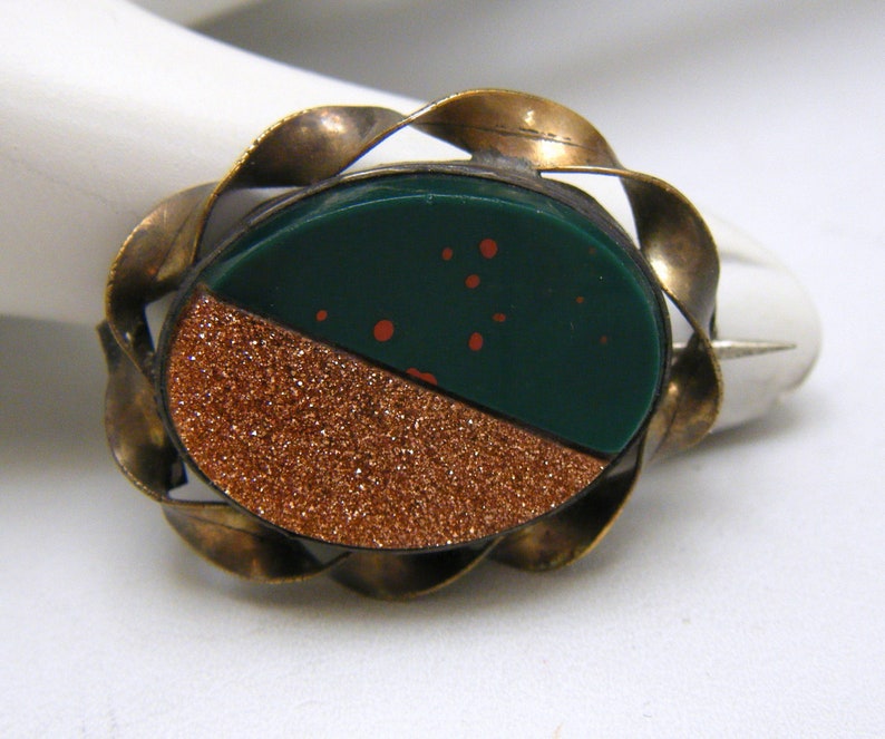 Puede incluir: Un broche vintage con una piedra de color verde y cobre engastada en un marco de lat&oacute;n. La piedra est&aacute; dividida en dos, un lado verde oscuro con motas naranjas y el otro lado un brillo de color cobre.