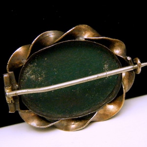 Puede incluir: Un broche de tono plateado y dorado con un camafeo verde. El broche tiene un cierre de pin y enganche.