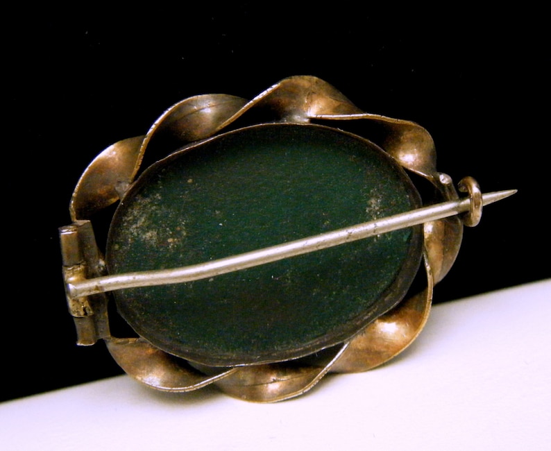 Puede incluir: Un broche de tono plateado y dorado con un camafeo verde. El broche tiene un cierre de pin y enganche.