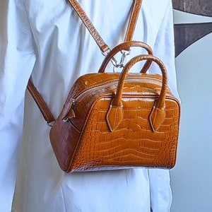 Puede incluir: Una mochila estructurada marrón con textura de piel de cocodrilo. La bolsa tiene un asa superior y correas ajustables. La cremallera y los herrajes son plateados. La bolsa se lleva sobre un vestido blanco.