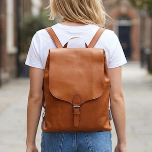 May include: A tan leather backpack with a large fold-over flap, a single vertical strap with a silver-toned metal buckle closure, and two shoulder straps, worn over a plain white t-shirt and blue denim jeans.