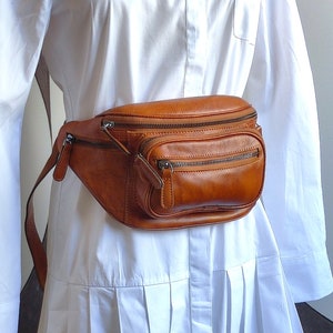 May include: A brown leather fanny pack with multiple zippered compartments. The bag is worn around the waist of a person wearing a white shirt.