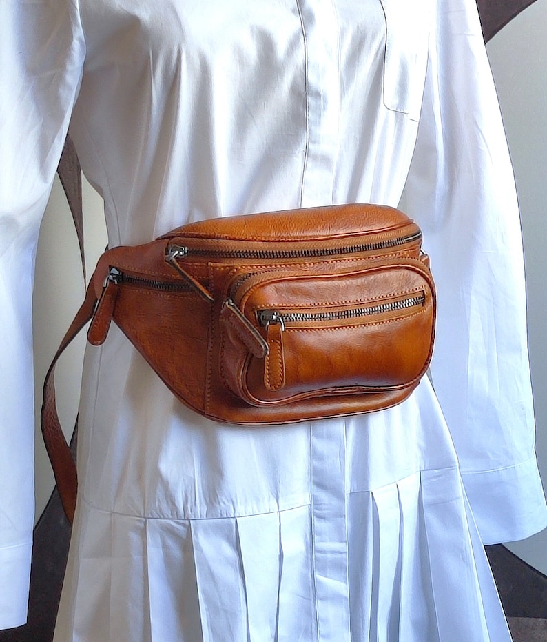 May include: A brown leather fanny pack with multiple zippered compartments. The bag is worn around the waist of a person wearing a white shirt.