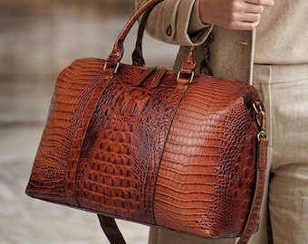 Könnte beinhalten: Eine große, rechteckige braune Reisetasche mit strukturiertem Krokodilledermuster. Die Tasche verfügt über zwei Tragegriffe, einen abnehmbaren Schulterriemen und goldfarbene Metallbeschläge. Die Oberfläche hat ein glänzendes Finish mit einem zentralen Prägemuster.