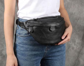 May include: A black leather fanny pack with a curved top zipper closure, a small front flap with a tab, and an adjustable strap. The bag features a smooth texture and a rounded silhouette, worn around the waist over a white t-shirt and blue jeans.