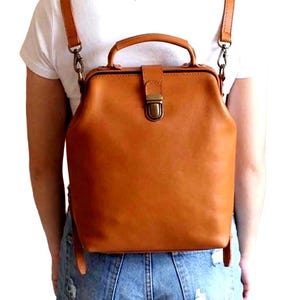 Peut inclure: Un sac à dos en cuir beige avec une structure rigide de type sac de médecin, doté d'une poignée supérieure, de bretelles réglables avec des clips en métal et d'une fermeture à rabat central avec un fermoir en métal doré.