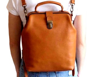 May include: A tan leather backpack with a structured doctor bag style frame, featuring a top handle, adjustable shoulder straps with metal clips, and a central front flap closure with a brass-toned metal clasp mechanism.