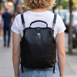 May include: A black structured backpack with a doctor-style frame opening, a top handle, and a central metal clasp closure. The bag features adjustable shoulder straps attached with metal hardware, providing a clean and functional design.