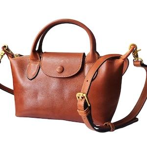 Könnte beinhalten: Eine braune Lederhandtasche mit einem oberen Griff und einem abnehmbaren Schulterriemen. Die Tasche hat einen Klappenverschluss mit einem Knopf und goldfarbener Hardware. Die Tasche ist etwa 25 cm breit.