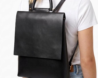 May include: A black backpack with a smooth finish, featuring a large rectangular flap closure, a rounded top handle, and adjustable shoulder straps. The bag has a structured, clean-lined design with metallic hardware accents at the handle base.