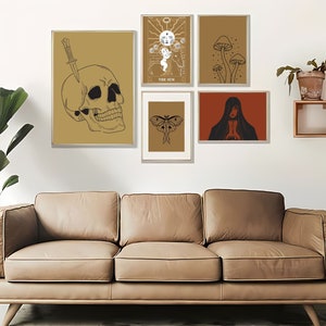 May include: A collection of framed art prints featuring mystical and gothic themes. The prints include a skull with a knife, tarot card imagery, mushrooms, a moth, and a cloaked figure. The color palette is primarily gold, brown, and black.