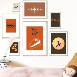 May include: A collection of framed Halloween-themed art prints. The prints feature orange, black, and white designs, including text that reads "Spooky", a coffin, a witch on a broom, and candy corn. The frames are gold.