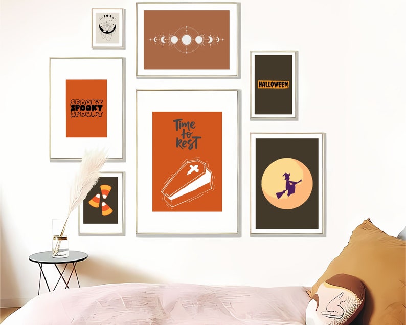 May include: A collection of framed Halloween-themed art prints. The prints feature orange, black, and white designs, including text that reads "Spooky", a coffin, a witch on a broom, and candy corn. The frames are gold.