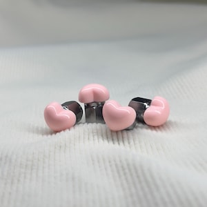 Könnte beinhalten: Eine Sammlung von rosa herzförmigen Dekorationsgegenständen mit einem metallischen Sockel. Die Herzen sind in einem weichen, pastellrosa gehalten. Die Metallsockel sind dunkelgrau. Die Objekte sind auf einer weißen, strukturierten Oberfläche angeordnet.