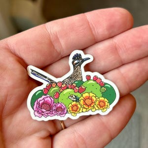 May include: A sticker featuring a cartoon roadrunner perched on a prickly pear cactus with pink and yellow flowers. The roadrunner is brown and black with a long tail and a pointed beak. The cactus is green with red berries.