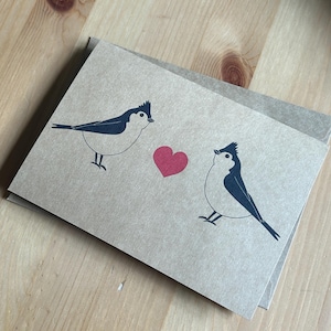 May include: A pair of greeting cards with a minimalist design featuring two stylized birds facing each other with a red heart between them. The birds are black and white on a tan background. The cards are on a wooden surface.