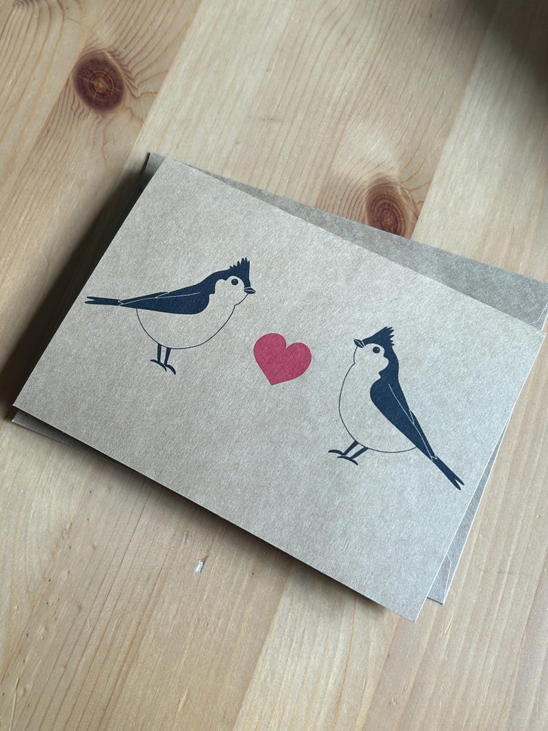May include: A pair of greeting cards with a minimalist design featuring two stylized birds facing each other with a red heart between them. The birds are black and white on a tan background. The cards are on a wooden surface.