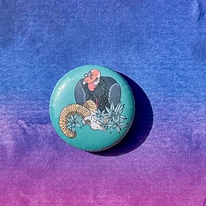 May include: A round button with a turquoise background featuring a black and red condor perched on a ram skull with green foliage.