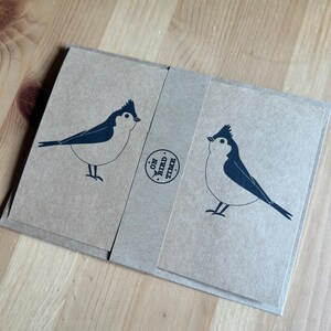 May include: A set of kraft paper cards with a dark blue bird illustration. The cards are wrapped with a matching paper band that has the text "ON BIRD TIME" printed on it. The bird design is simple and modern, with a minimalist aesthetic.