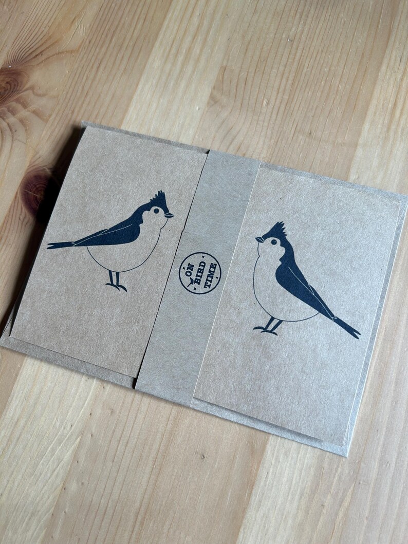 May include: A set of kraft paper cards with a dark blue bird illustration. The cards are wrapped with a matching paper band that has the text "ON BIRD TIME" printed on it. The bird design is simple and modern, with a minimalist aesthetic.