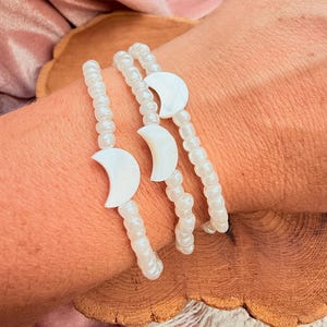 May include: Three beaded bracelets with white moon-shaped charms. The bracelets are made of small, iridescent beads. The moon charms are a bright white color. The bracelets are displayed on a wooden surface.