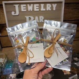 Puede incluir: Dos bolsas transparentes llenas de cuentas coloridas, atadas con hilo y peque&ntilde;as cucharas de madera. Cada bolsa tiene una etiqueta con el texto "dream" y una direcci&oacute;n web. El fondo presenta un letrero que dice "JEWELRY BAR".