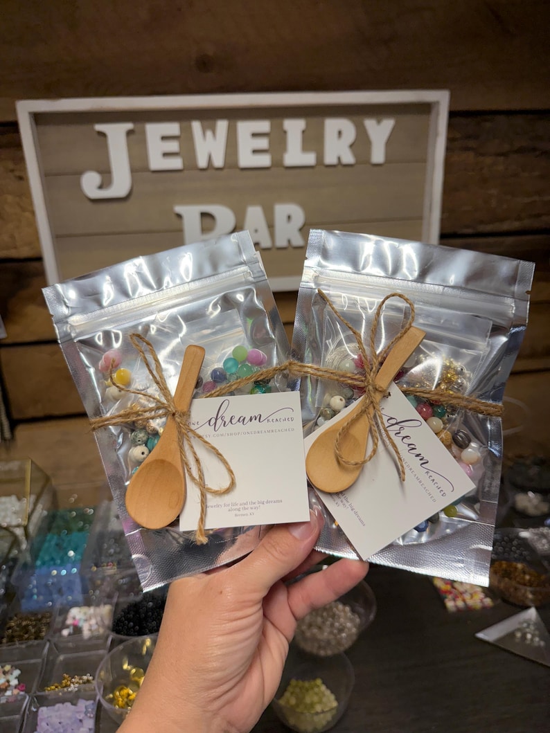 Puede incluir: Dos bolsas transparentes llenas de cuentas coloridas, atadas con hilo y peque&ntilde;as cucharas de madera. Cada bolsa tiene una etiqueta con el texto "dream" y una direcci&oacute;n web. El fondo presenta un letrero que dice "JEWELRY BAR".