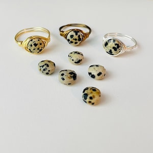 May include: Three wire-wrapped rings with dalmatian jasper stones, alongside loose stones. The rings feature gold and silver wire. The dalmatian jasper stones are beige with black spots, creating a unique and natural look. Jewelry.